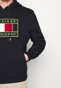 Felpa con cappuccio blu scuro che presenta un grande logo ricamato con rifiniture verdi, rosse, bianche e dorate. Realizzata in misto cotone, dotata di tasca a marsupio e cordini regolabili.