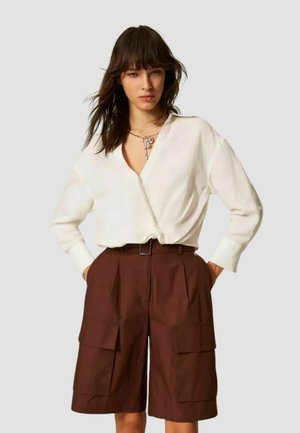 Donna che indossa una blusa bianca a maniche lunghe infilata in pantaloncini cargo marroni a vita alta e lunghezza al ginocchio con cintura, mani nelle tasche.