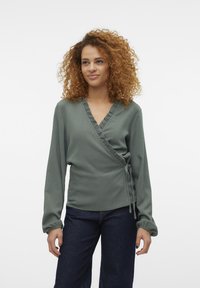 Blouse verte avec un tissu texturé, col volanté et manches longues, assortie à un jean en denim foncé. Lien ajustable à la taille.
