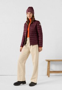 Chaqueta acolchada burdeos de corte ajustado, suéter de cuello alto naranja, pantalones crema y gorro burdeos acanalado. Zapatos negros sin cordones y banco de madera.