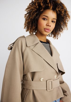 Noor naine lokkis juustega, kandes beežikat vöölõikega trenškihti must-valge triibulise särgi kohal tavalise tausta ees.