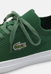 Sapatilha em malha verde com tecido texturizado, atacadores planos, sola em borracha e um detalhe branco. Apresenta um pequeno logo de crocodilo bordado.