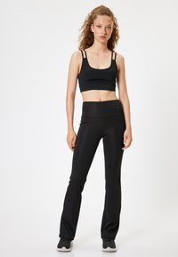 Sujetador deportivo negro con tirantes finos, combinado con leggings negros acampanados de cintura alta. Ambos están hechos de material elástico. Zapatillas grises completan el look.