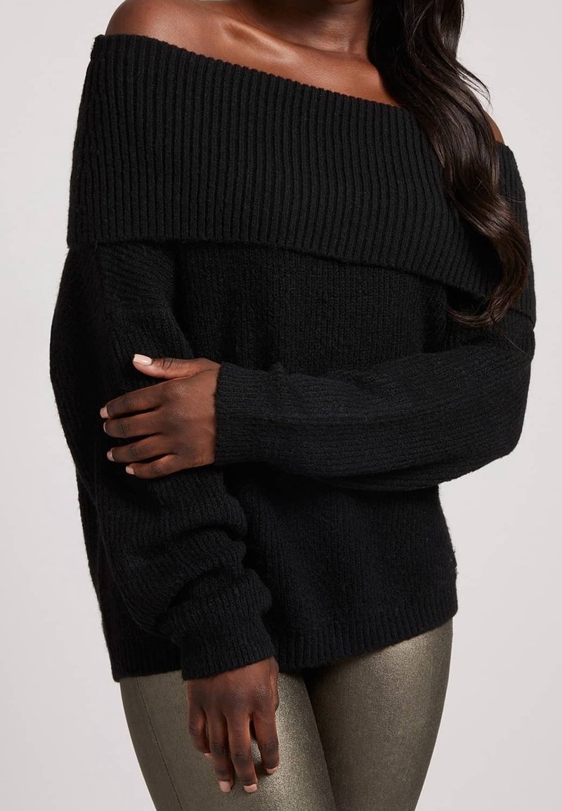 Femme portant un pull noir côtelé à col bateau et manches longues, avec un pantalon argenté métallisé ajusté, sur fond uni.