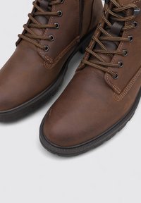 Botas de cuero marrón con suelas de goma negra, puntera redonda y cordones redondos. Presentan detalles de costura y ojales metálicos para los cordones.