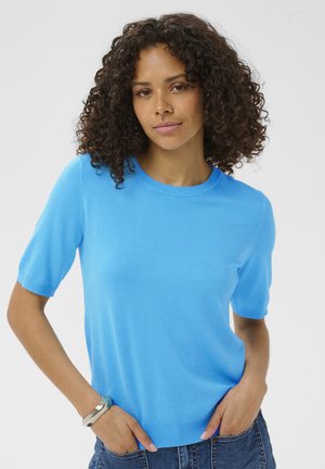 Femme aux cheveux bouclés portant un pull à manches courtes bleu vif et un jean, debout les mains dans les poches devant un fond uni.