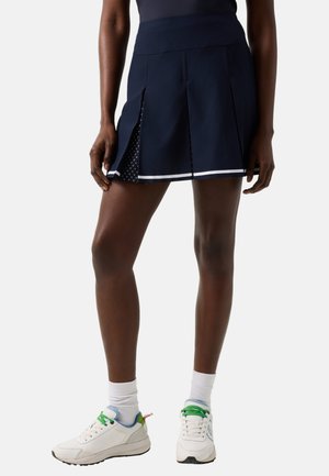 Kvinde iført en navy plisseret nederdel med hvid kant og hvide sneakers med grønne snørebånd, stående mod en ensfarvet baggrund.