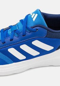 Chaussure de sport bleue avec un dessus en maille et synthétique, design à trois rayures blanches, languette texturée, lacets plats et semelle en caoutchouc blanche.
