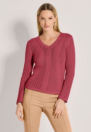 Vrouw met steil blond haar, gekleed in een roze kabelgebreide V-halstrui en beige broek, staand tegen een effen lichte achtergrond.