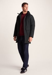 Man wearing a black hooded coat, burgundy sweater, dark jeans, and black sneakers standing against a plain light background.