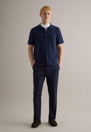 Hombre joven de pie con las manos en los bolsillos, vistiendo una camisa azul marino de manga corta abotonada sobre una camiseta blanca, pantalones oscuros y zapatos marrones.