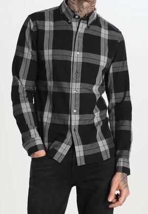 Mann trägt ein großes schwarz-grau kariertes Hemd mit Knöpfen und Kragen, schwarze Hose und sichtbare Tätowierungen am Hals und an den Händen.