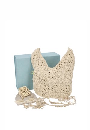 Bolso de hombro de ganchillo color crema, expuesto con accesorios de borlas a juego y joyas de cuentas de madera, delante de una caja azul claro.
