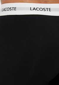 Fekete boxer alsónadrág fehér gumipánttal, amelyen fekete színnel a "LACOSTE" felirat látható. Az anyag puhanak és rugalmasnak tűnik.