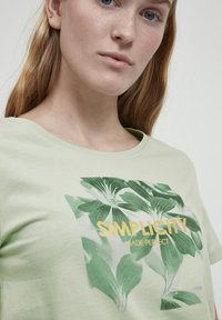 Camiseta de algodón verde claro con un estampado floral y el texto "Simplicidad Hecha Perfecta" en amarillo. Diseño de cuello redondo.
