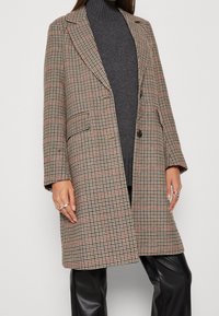 Manteau en mélange de laine à carreaux avec un col cranté, deux boutons et des poches. Tons neutres avec des accents rouges. Associé à un col roulé gris et un pantalon noir.