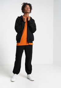 Sudadera negra con cremallera sobre una camiseta naranja, combinada con pantalones de chándal negros y zapatillas blancas, sobre un fondo claro y liso.