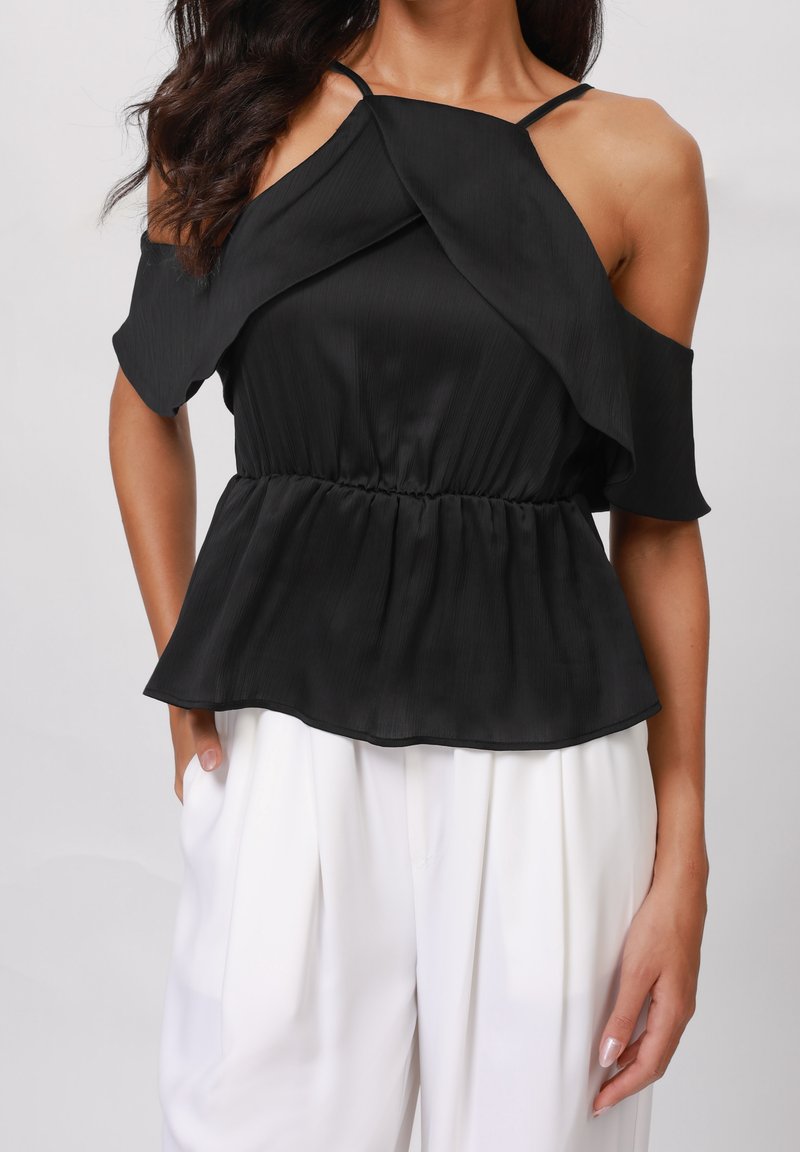 Blouse noire épaules dénudées avec plis, taille élastique et fines bretelles. Texture lisse et design fluide complètent un pantalon blanc.