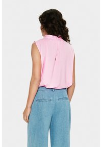 Różowa bluzka bez rękawów z marszczonym kołnierzem i detalem z guzikami z tyłu, zestawiona z jasnoniebieskimi spodniami o szerokich nogawkach z tylnymi kieszeniami.