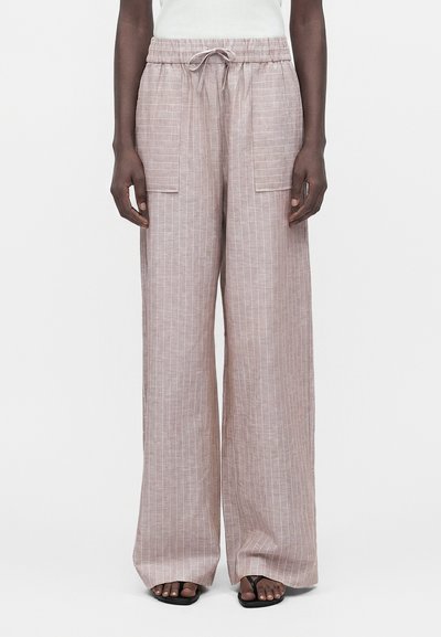 Pantalon large en lin beige avec des rayures verticales blanches, taille élastique, poches plaquées devant, porté avec des sandales noires et un top blanc.