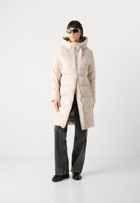 Beige pufferjas met een hoge kraag, ritssluiting en elastische accenten. Draagt over donkergrijze wijde broek en zwarte schoenen.