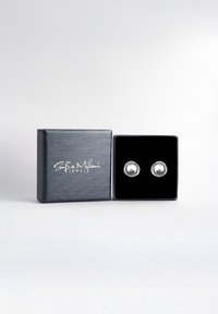 Silverfärgade studsörhängen inuti en svart sammetsbehållare, placerade i en texturerad svart låda med en vit logotyp. Rund form med en slät yta.