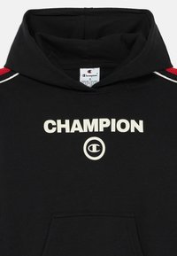Fleece preto da Champion com texto branco a dizer "CHAMPION" e logótipo. Apresenta pormenores em vermelho nos ombros e um bolso frontal. Tecido suave.