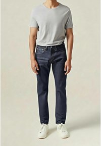 Mörkblå jeans med smal passform, fem fickor och kontrasterande sömmar, kombinerade med en ljusgrå kortärmad skjorta och vita sneakers.