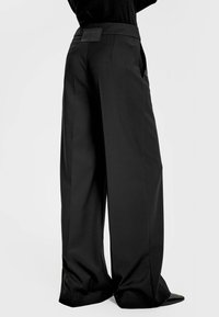 Czarne spodnie o szerokich nogawkach z gładkiego materiału, wysoki stan i detal z metką na plecach. Charakteryzują się czystym, dopasowanym wyglądem i swobodnym krojem.