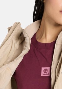 Chaqueta beige con cremallera y un suave interior de forro polar, superpuesta a una camiseta de color marrón profundo con un logo de Timberland en rosa.