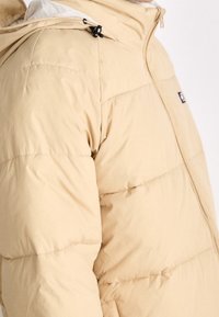 Chaqueta acolchada beige con textura suave, grandes secciones acolchadas y cierre frontal con cremallera. Cuenta con una capucha y un pequeño logo en el pecho.