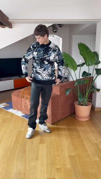 Jovem ajustando suéter com padrão preto, branco e azul, em pé sobre o chão de madeira perto de um sofá em tom ferrugem e de uma grande planta em vaso na sala de estar.