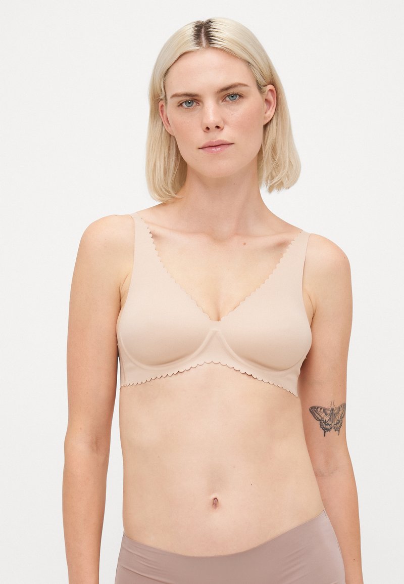 Beige bra with scalloped edges, smooth fabric, and molded cups. Wide straps for support, featuring a simple, seamless design.