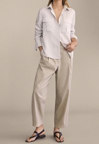 Lekka, biała koszula z guzikami, z dwiema kieszeniami na piersi, w połączeniu z beżowymi spodniami o szerokich nogawkach i czarnymi sandałami na ramiączkach na neutralnym tle.