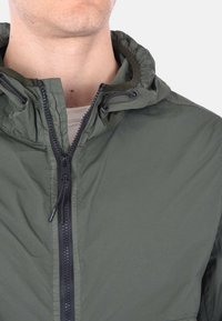 Giacca leggera verde oliva con cappuccio, dotata di zip nera e cordino regolabile. Presenta una texture liscia e un design minimalista.