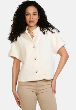 Frau mit langen lockigen Haaren, die eine kurzärmelige, geknöpfte, elfenbeinfarbene Jacke und beige Hose trägt und mit einer Hand auf der Hüfte steht.