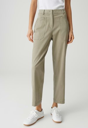 Persoon die beige rechte broek met voorzakken en witte sneakers draagt, staand tegen een neutrale achtergrond.