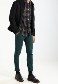 Svart ullkappa med en fårskinnskrage, rutigt knopplåtskjorta, gröna slim-fit byxor och svarta snörskor.