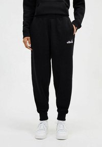 Pantalones de chándal negros con cintura y puños elásticos, que presentan un logo blanco en la pierna izquierda. Se combinan con zapatillas blancas.