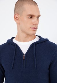 Mörkblå zippehoodie gjord av texturerat tyg, med en snoddhuva och en kontrasterande vit t-shirt under.
