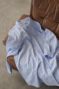 Chemise bleu clair à manches longues et boutons, posée sur le bras et l'assise d'une chaise en cuir marron sur un sol en béton.