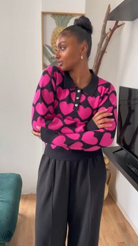 Roza pulover s vzorcem srčkov na črni podlagi, z ovratnikom in tremi gumbi v obliki srčkov. Kombiniran z sproščenimi črnimi hlačami.