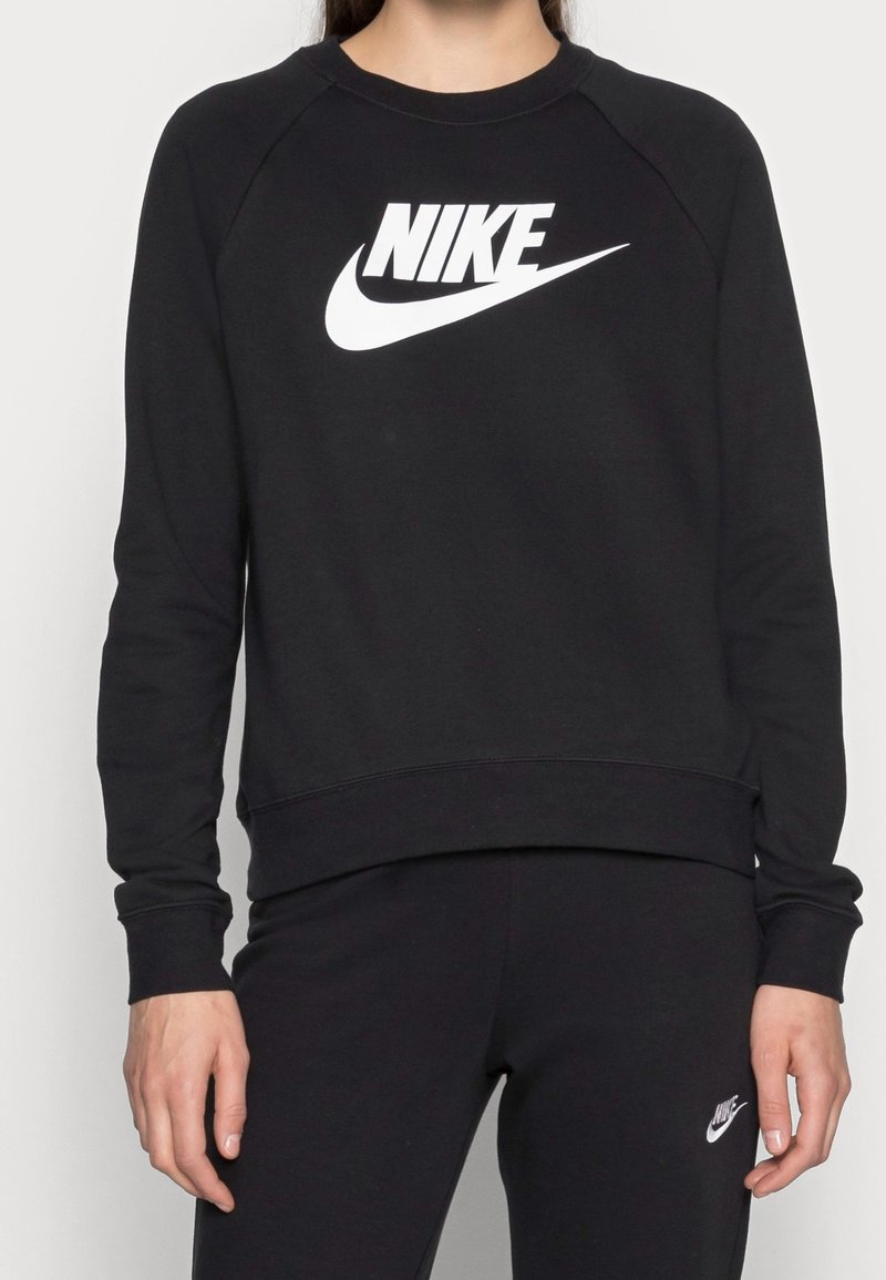 Person wearing black Nike sweatshirt and black pants, both with white Nike logo on front, standing against a plain background.