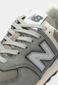 Sneaker New Balance grigia e beige con lacci bianchi, logo cucito di lato e etichetta con marchio sulla linguetta in vista ravvicinata.