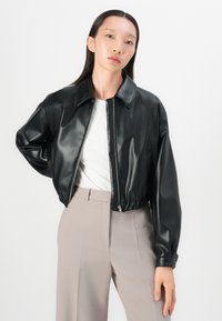 Giacca cropped in ecopelle nera con colletto piatto, chiusura con zip e vita elasticizzata. Il modello indossa una maglietta bianca e pantaloni beige a vita alta.