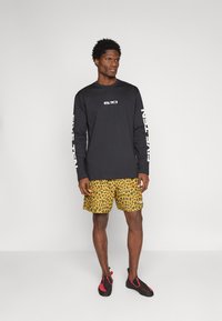 Črna majica z dolgimi rokavi z belim napisom, kombinirana s svetlo rumenimi kratkimi hlačami s potiskom leopardjega vzorca. Model nosi črne plezalne čevlje.