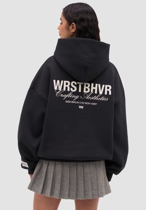 Femme aux cheveux foncés portant un sweat à capuche noir oversize avec un texte blanc et une jupe plissée grise, vue de profil sur un fond uni.