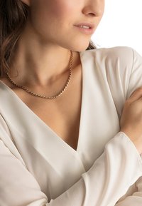 Gold-toned chain necklace with a twisted design, worn against a light-colored blouse featuring a V-neck and flowing sleeves.