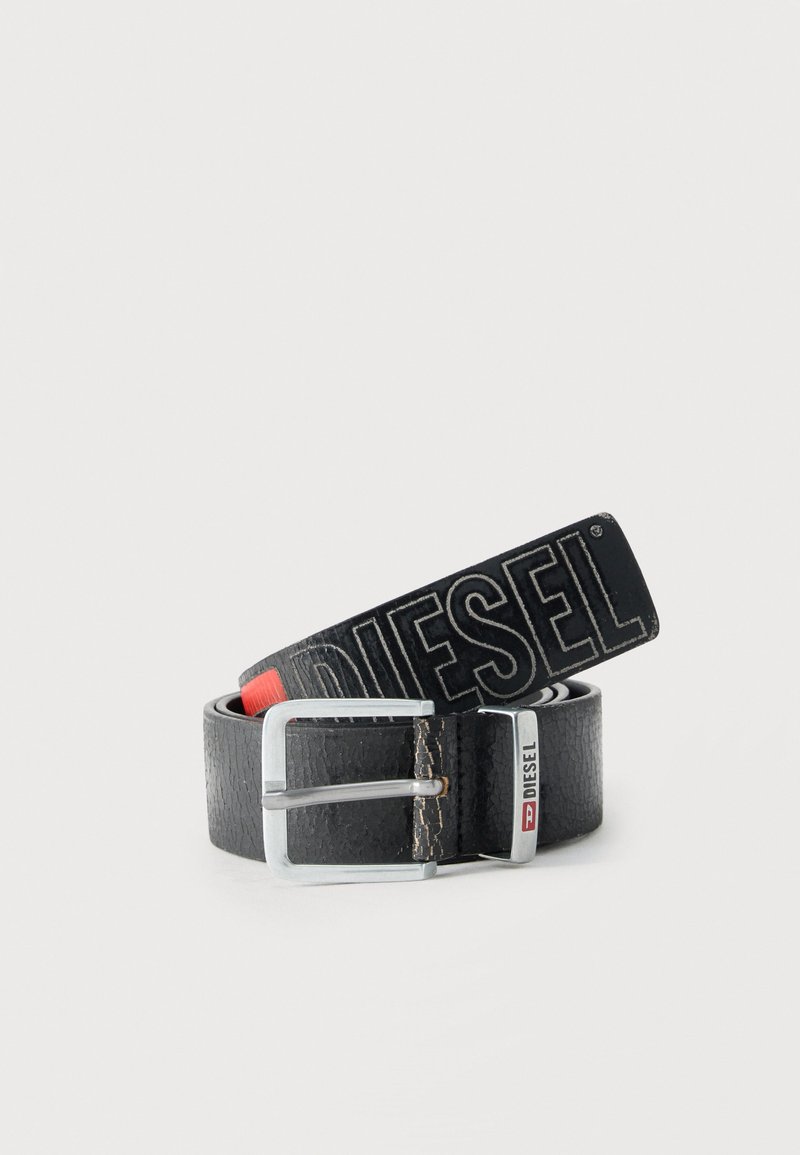 Μαύρη ζώνη από δερματόυχο με υφή, με ασημένια πόρπη και την επιγραφή "DIESEL" σε μεγάλα γράμματα κατά μήκος του ιμάντα, καθώς και μικρή μεταλλική ετικέτα στη θηλιά.