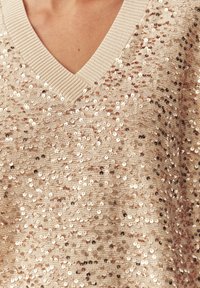 Bēša top ar spīdīgiem fliteriem, V-veida izgriezumu un ribotu apkakli. Audumam ir teksturēta virsma ar spožiem apaļiem fliteriem, kas vienmērīgi izvietoti.